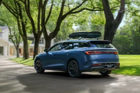 Blauer BYD SEAL 6 DM-i Touring mit Dachbox in seitlicher Heckansicht fährt auf ein Haus zu.