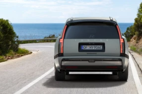 Die Heckansicht eines hellgrauen SUV, das auf einer kurvigen Straße am Meer entlangfährt.