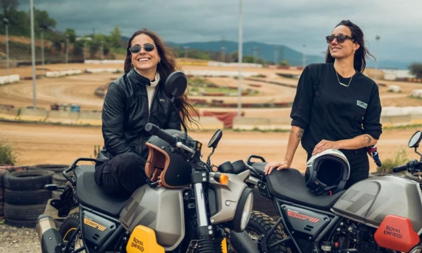 Zwei lächelnde Frauen sitzen auf ihren Motorrädern vor einer Fahrstrecke aus Sand.