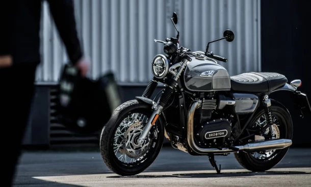 Ein Brixton Motorrad steht auf einem asphaltierten Vorplatz in einem Industriegebiet, während eine Person mit einem Helm in der Hand auf das Bike zusteuert.