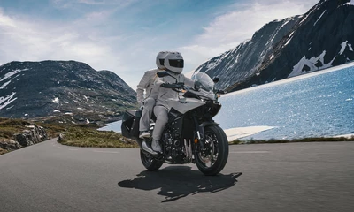 Zwei Personen fahren auf einem Motorrad vor einem See und Bergpanorama.