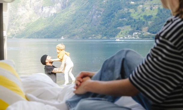 Eine Person sitzt im Inneren eines Wohnmobils und blickt auf einen Mann und ein Kleinkind, die am Ufer eines Sees spielen.