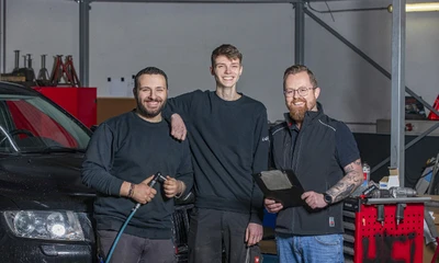 Drei Kummich Werkstatt-Mechatroniker stehen in der Werkstatt in Arbeitskleidung - bereit für Ihr Fahrzeug.