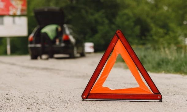 Ein aufgestelltes Unfalldreieck auf einem Schotterweg. Im Hintergrund einparkenedes Auto mit offener Heckklappe und eine Person in Warnweste.